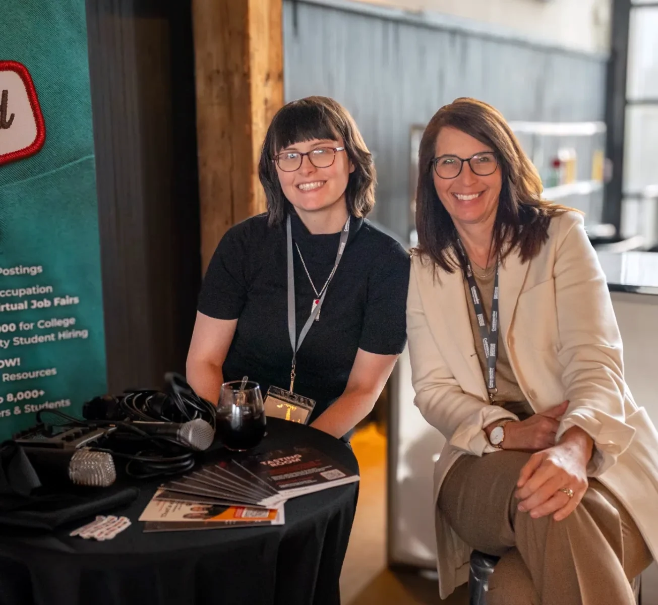 Two woman sitting a table with a camera and poster in the background.